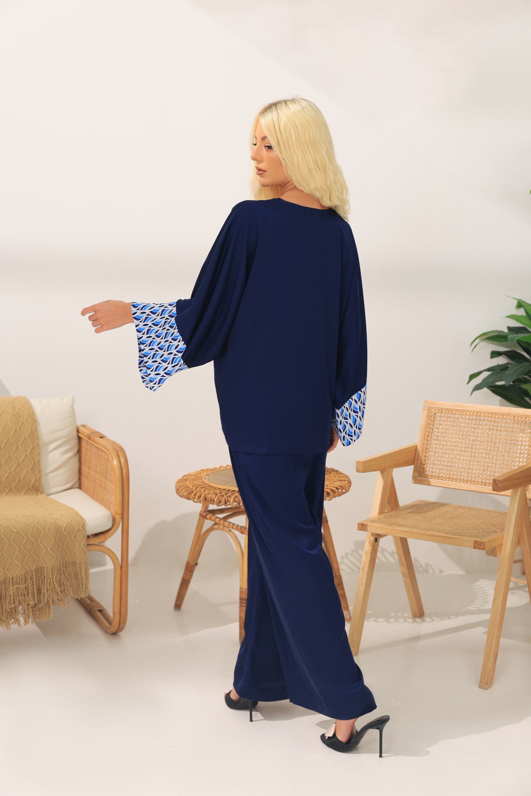 Woman in a navy outfit with Greece patterned sleeves standing in a room with wooden chairs and a plant.